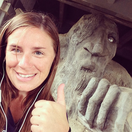 One of the few self-portraits, a woman holds her camera, smiles, and flashes the 'thumbs up' sign in front of one of geocaching's most famous landmarks (a giant troll scultpture in Seattle).