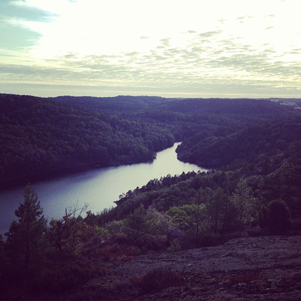 A bright sky with high clouds reflects on a faraway lake nestled among evergreens on all sides.