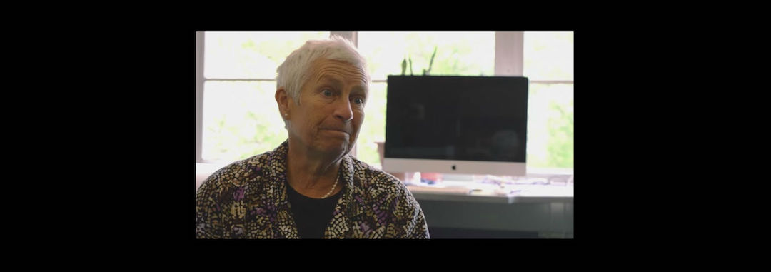 Cindy wearing a multi-colored shirt looking off camera, with an iMac on a desk behind her