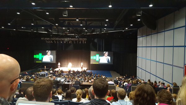 from the back of a large auditorium, a view of the stage with three small figures on it, flanked by two screens with Joyce's face and green bars on them