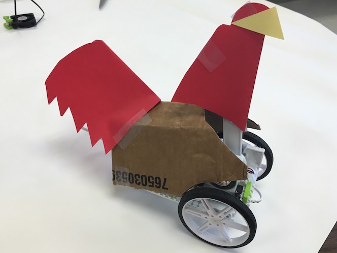 a littleBits construction of a chicken: over a littleBits invention (with two visible wheels) are a cardboard body and red and yellow construction paper for a tail and head