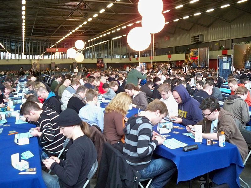 Dozens of Magic players sit at long tables in a large hall at a competitive Grand Prix tournament.