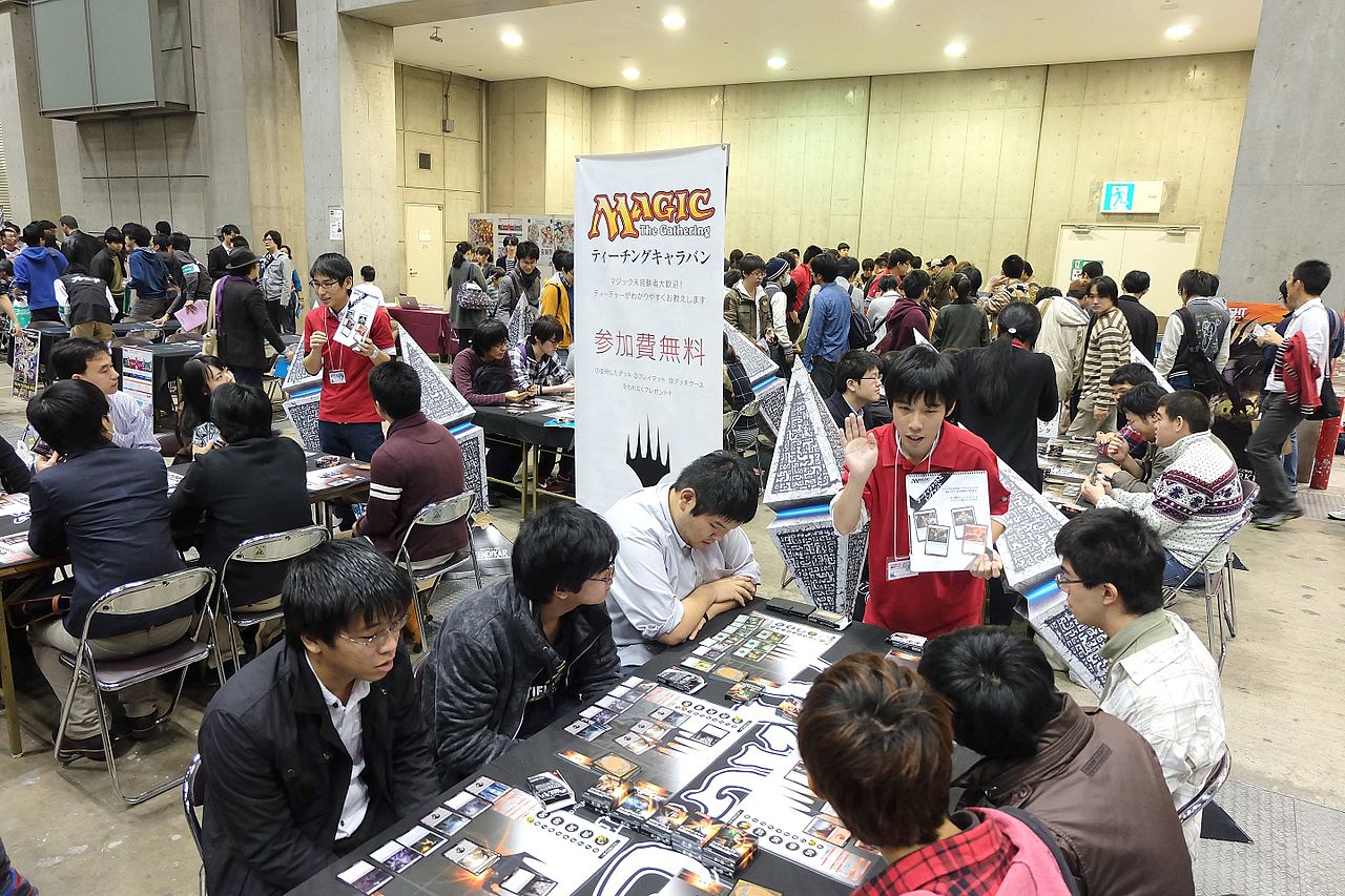 Two volunteers explain the basics of Magic to several new players seated at a gaming event in Tokyo.