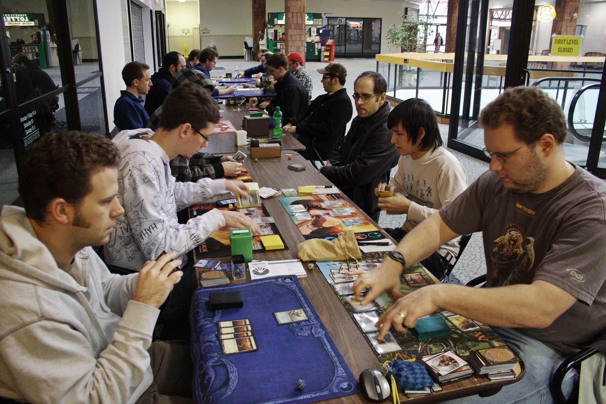 A photo of multiple Magic games occurring at once at a long row of tables outside a storefront in a mall.