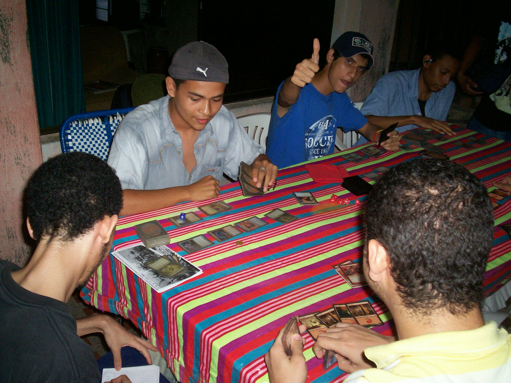 Several individuals sit at a table playing Magic. One of the players facing the camera holds out his hand in a 'thumbs-up' gesture.