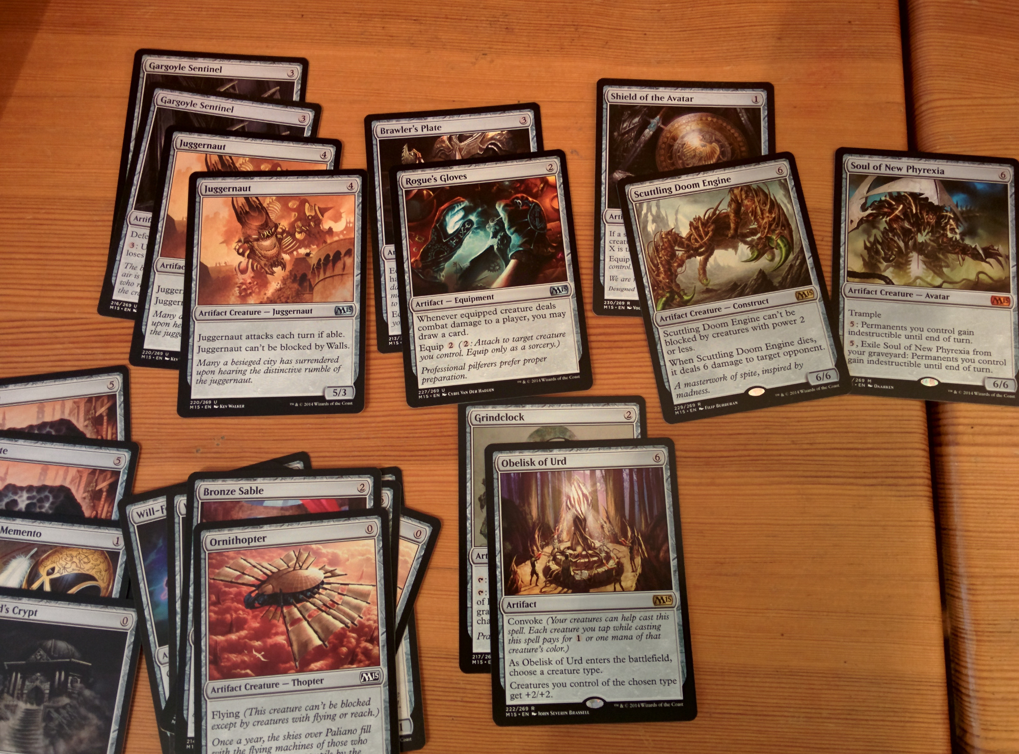 A number of Magic cards of the type 'Artifact' are spread out on a table surface, some stacked loosely on others. Cards include 'Juggernaut,' 'Rogue's Gloves,' and 'Obelisk of Urd.''