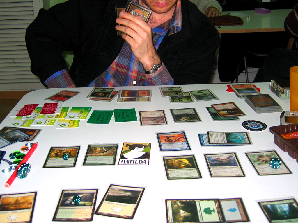 A game of Magic in play, focused on the cards controlled by one of the game's players. The opponent looks on from the opposite side of the table, sitting behind their own cards. Various cards controlled by each player are tapped.