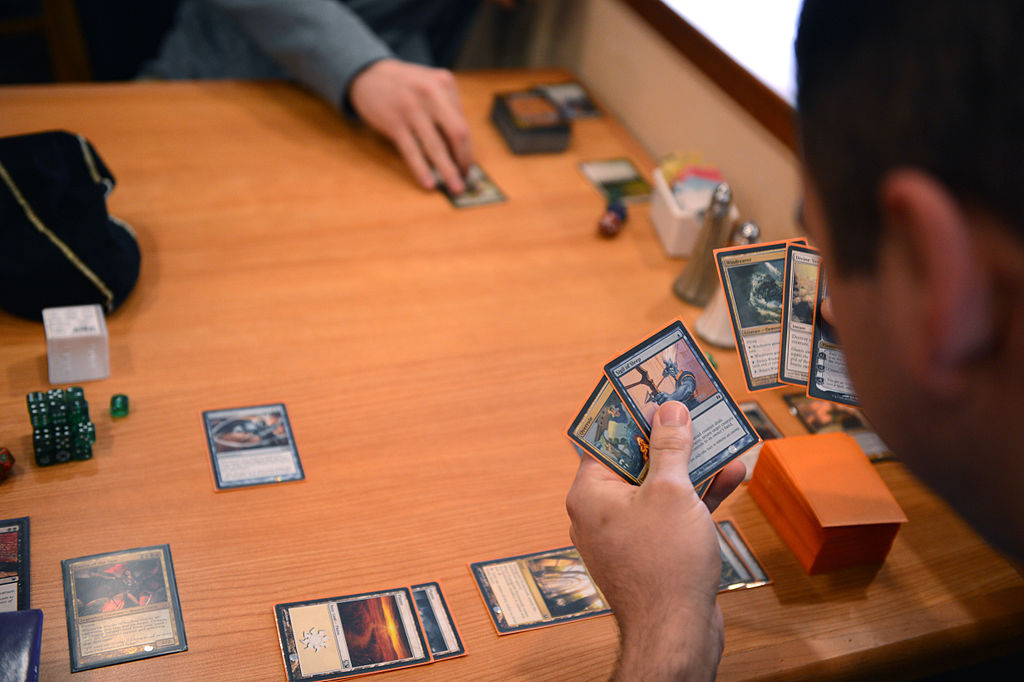 Two players engage in a game of Magic: The Gathering. One player reaches for a card on the table while the other looks through the cards in their hand.