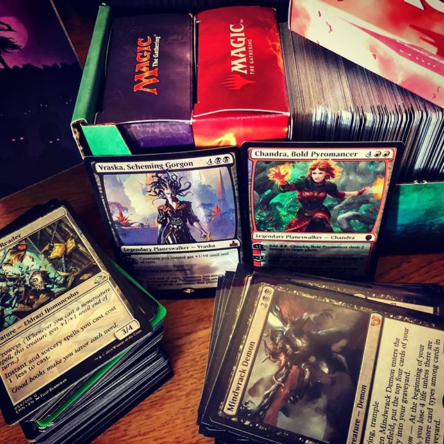 Two piles of Magic cards sit in front of two planeswalker cards leaning against a box filled with cards and deck boxes.