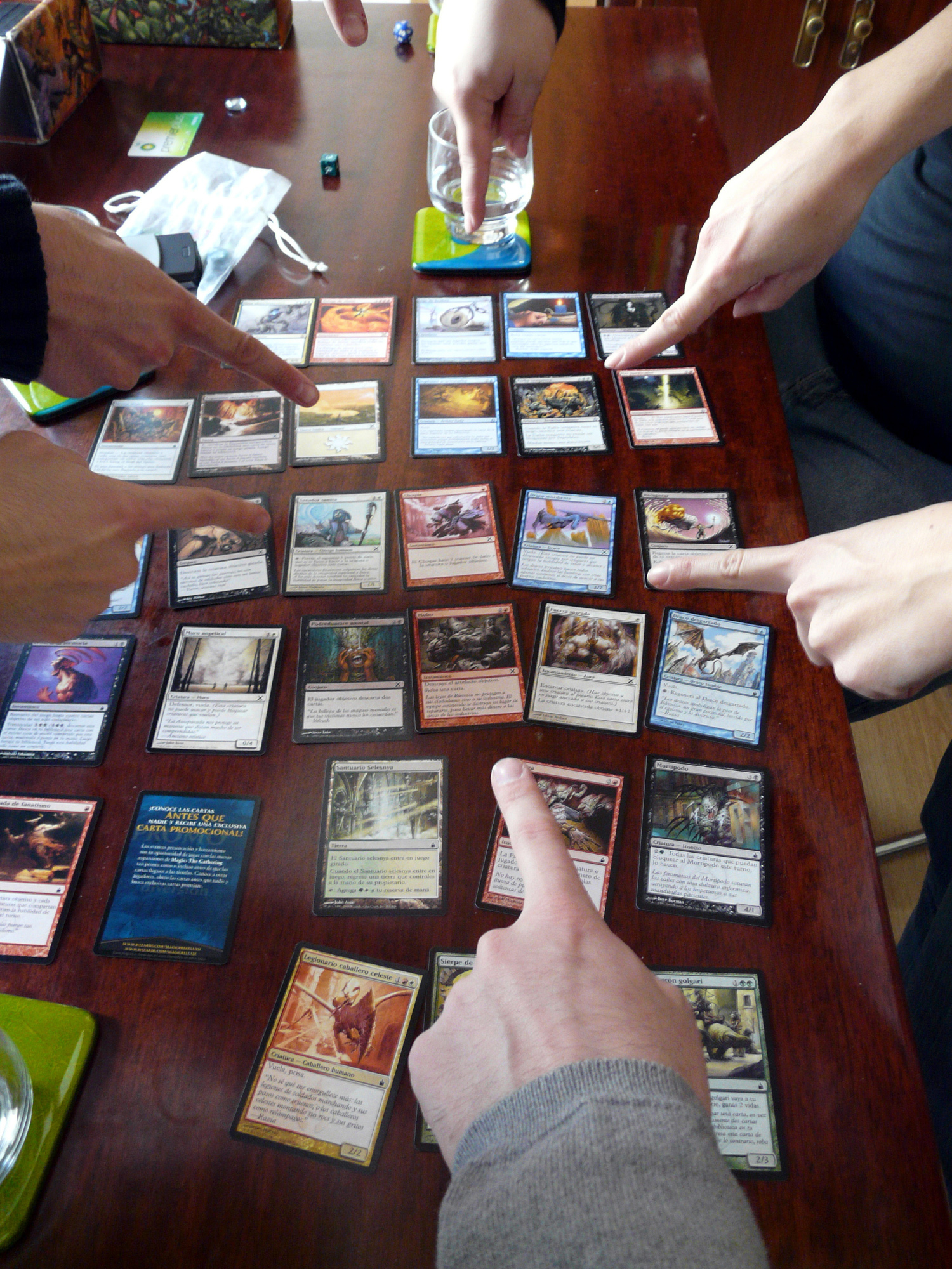Several hands from different edges of the photo point toward the center of the frame, in which a number of Magic cards are laid out in rows on a table.