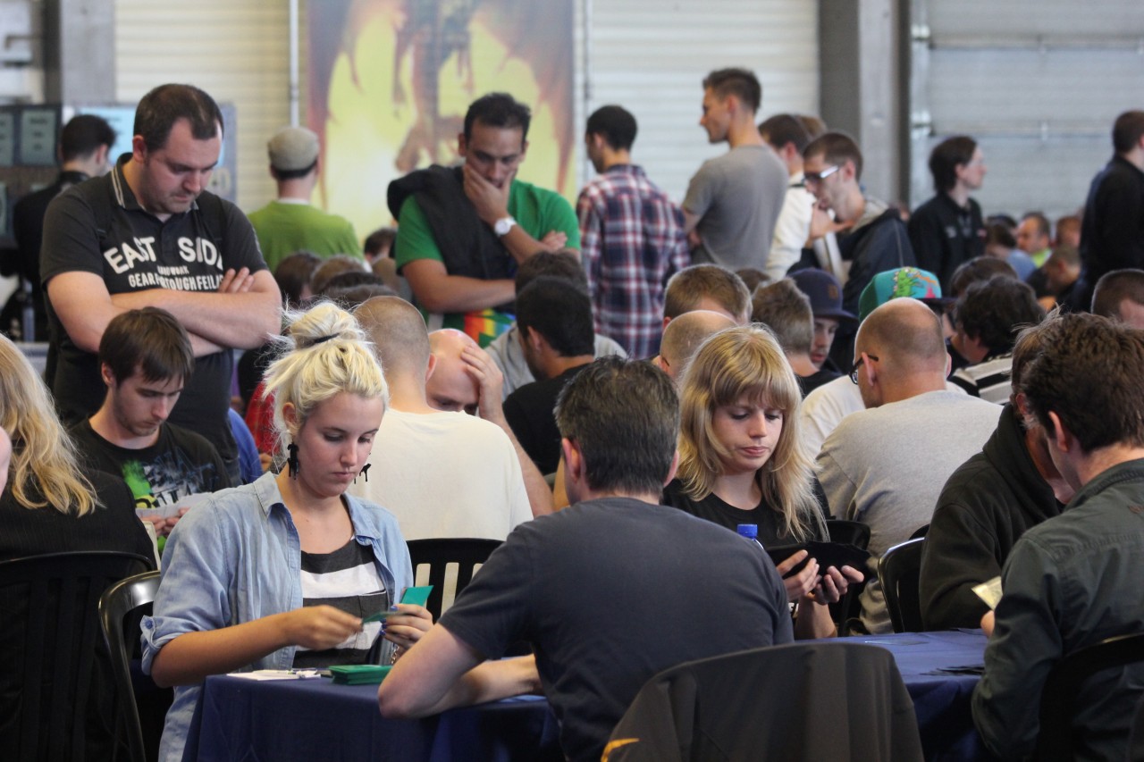 A number of Magic players are seated at tables to play in a competitive tournament. Some observers stand nearby to watch games in play.