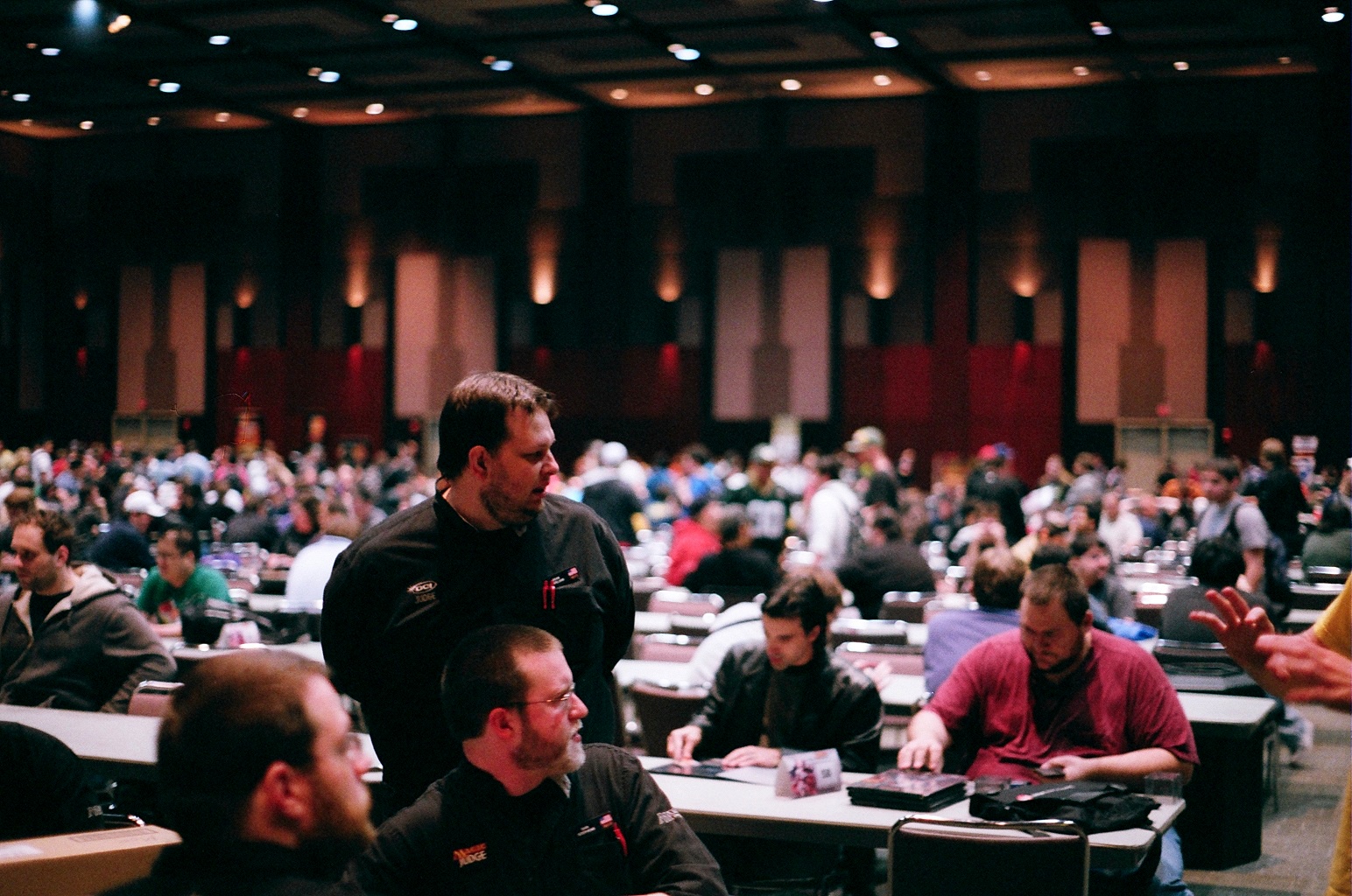 Several Magic judges, dressed in matching shirts noting their role, listen to a question or concern from a player at a competitive tournament.