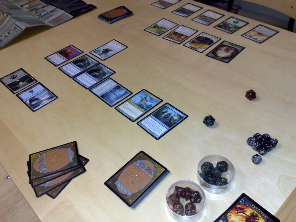 A photo of a Magic game 'battlefield' on which two players' cards are deployed. Visible cards include representations of creatures, mana-producing lands, and various enchantments and artifacts.