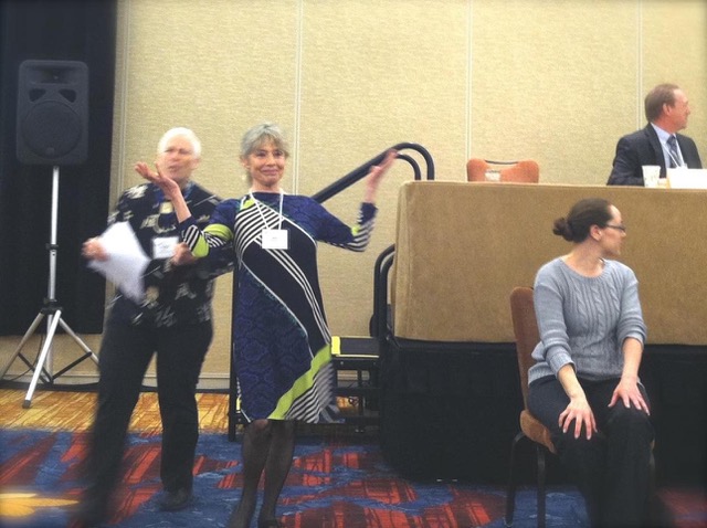 somewhat blurry photo of Gail in blue dress, in front of podium, arms raised; Cindy behind her, blurry