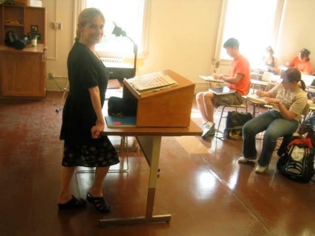 woman at podium in classroom, smiling at camera, with students in desks in front of her