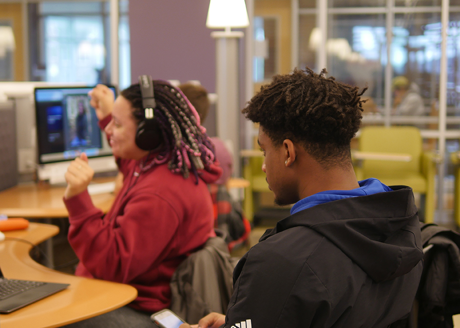 Student, seated at computer, using headphones and jamming out, raising arms like dancing or celebrating