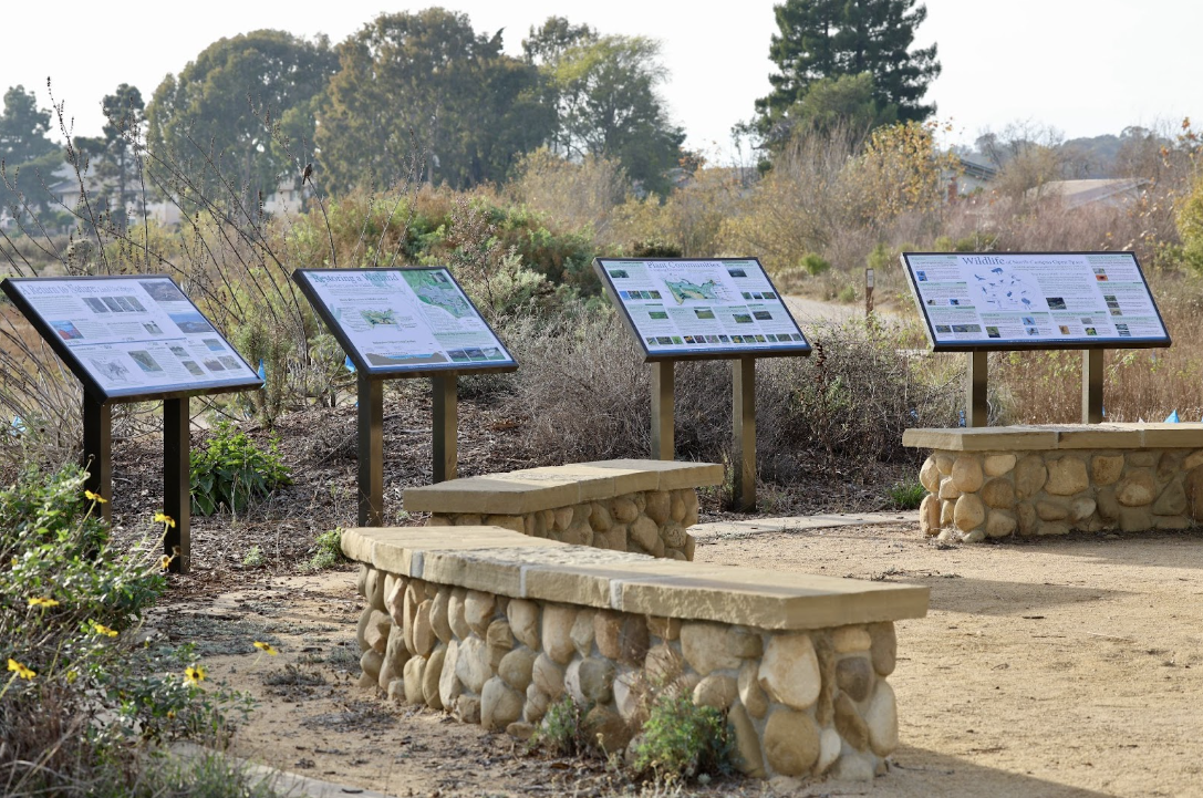 a restored estuary. In the foreground, there are four interpretive signs regarding the local ecology and three curved stone benches that form an outdoor classroom.
