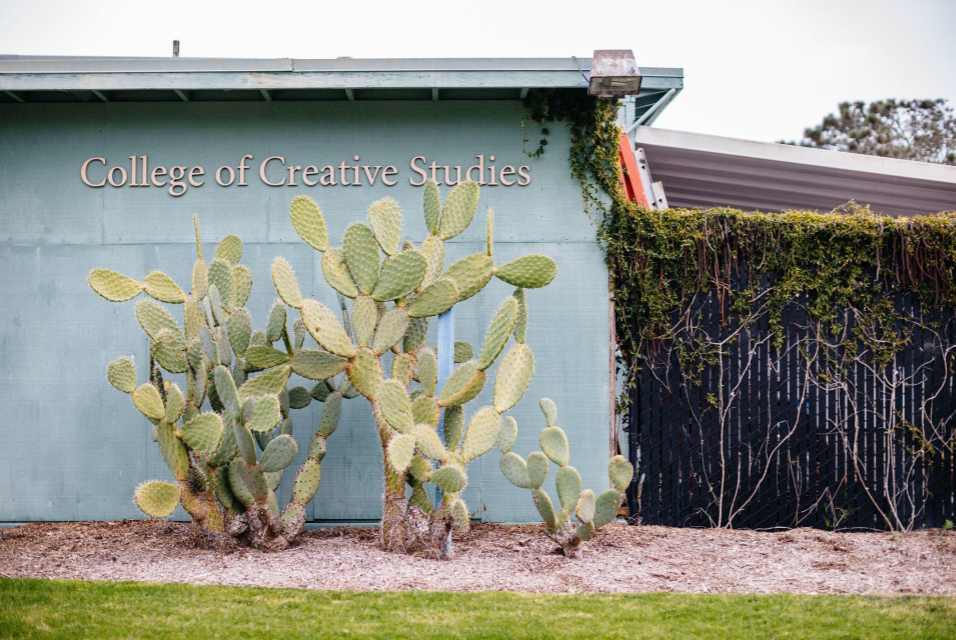 A faded green building with the name 'College of Creative Studies.' To the right of the building, there is a fence covered in shrubs. In front of the building, a large cactus plant partially obscures the building's name.