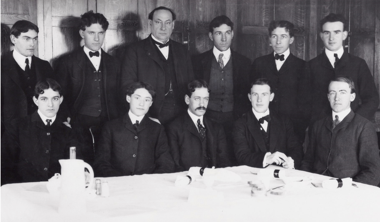 A black and white image in which Dr. Harvey Wiley and 10 of the volunteer male test subjects he enlisted into the so-called 'Poison Squad' experiments. The subjects are positioned in two rows, with those in the front row seated at a dining table and those in the back row, including Dr. Wiley, third from left, in a standing position. The subjects in the photograph are wearing suits and ties and facing forward, looking in front of them or at the camera, with calm or serious expressions.
