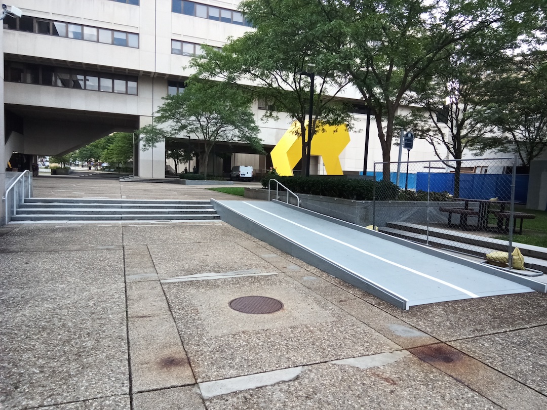 A set of stairs next to a metal ramp in an empty courtyard flanked by tall buildings.