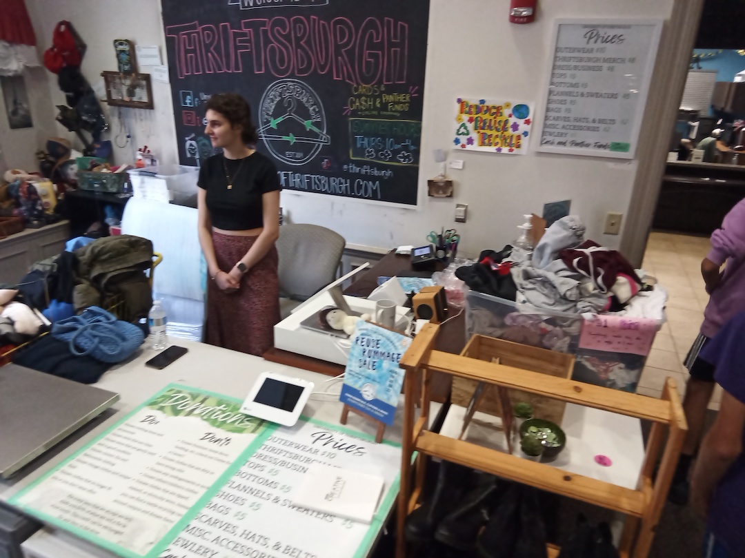 Woman standing behind the desk in the re-use clothing shop. A sign hanging behind her reads, Thriftsburgh. 
    