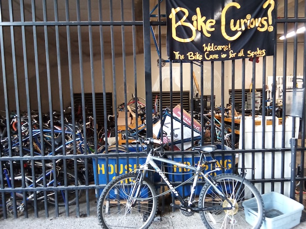 A bicycle leaning against a black fence with a sign that says, Bike Curious hanging above it.