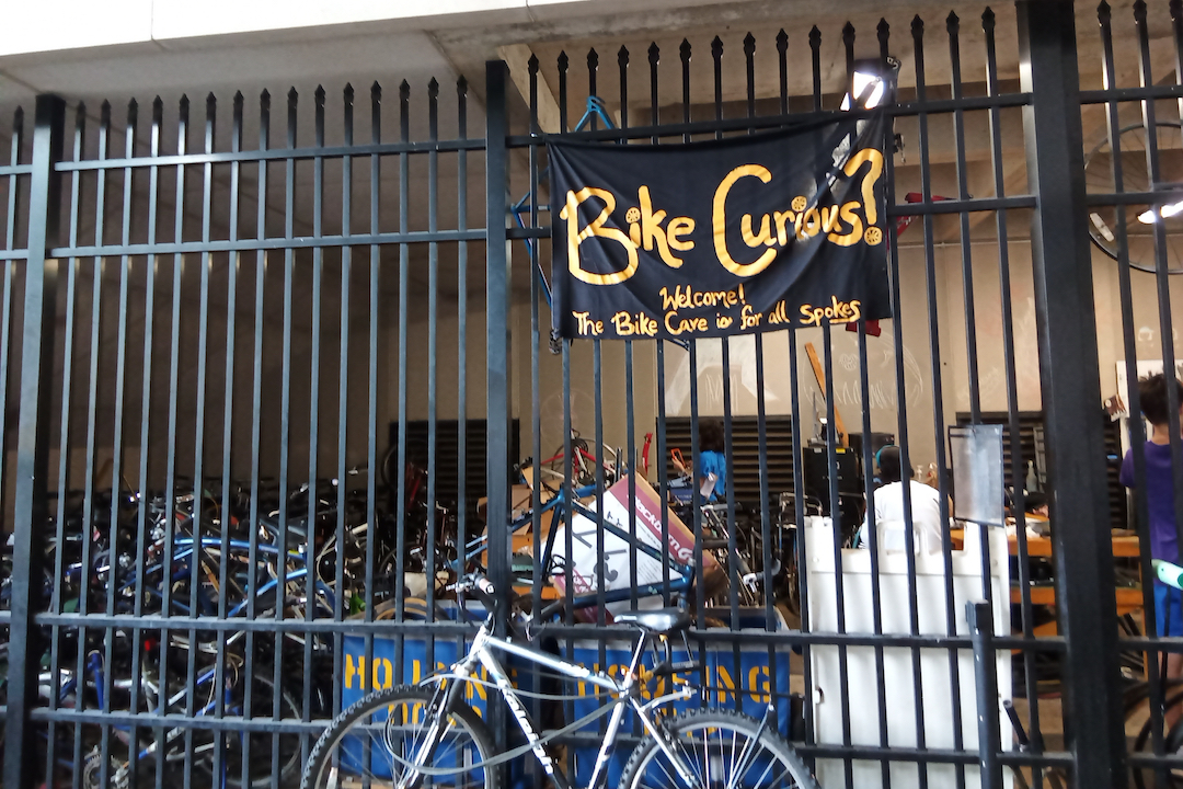 A bicycle leaning against a large black fence with a sign that says, Bike Curious hanging above it.