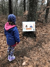 little girl standing in the woods, facing a sign with text and visuals.