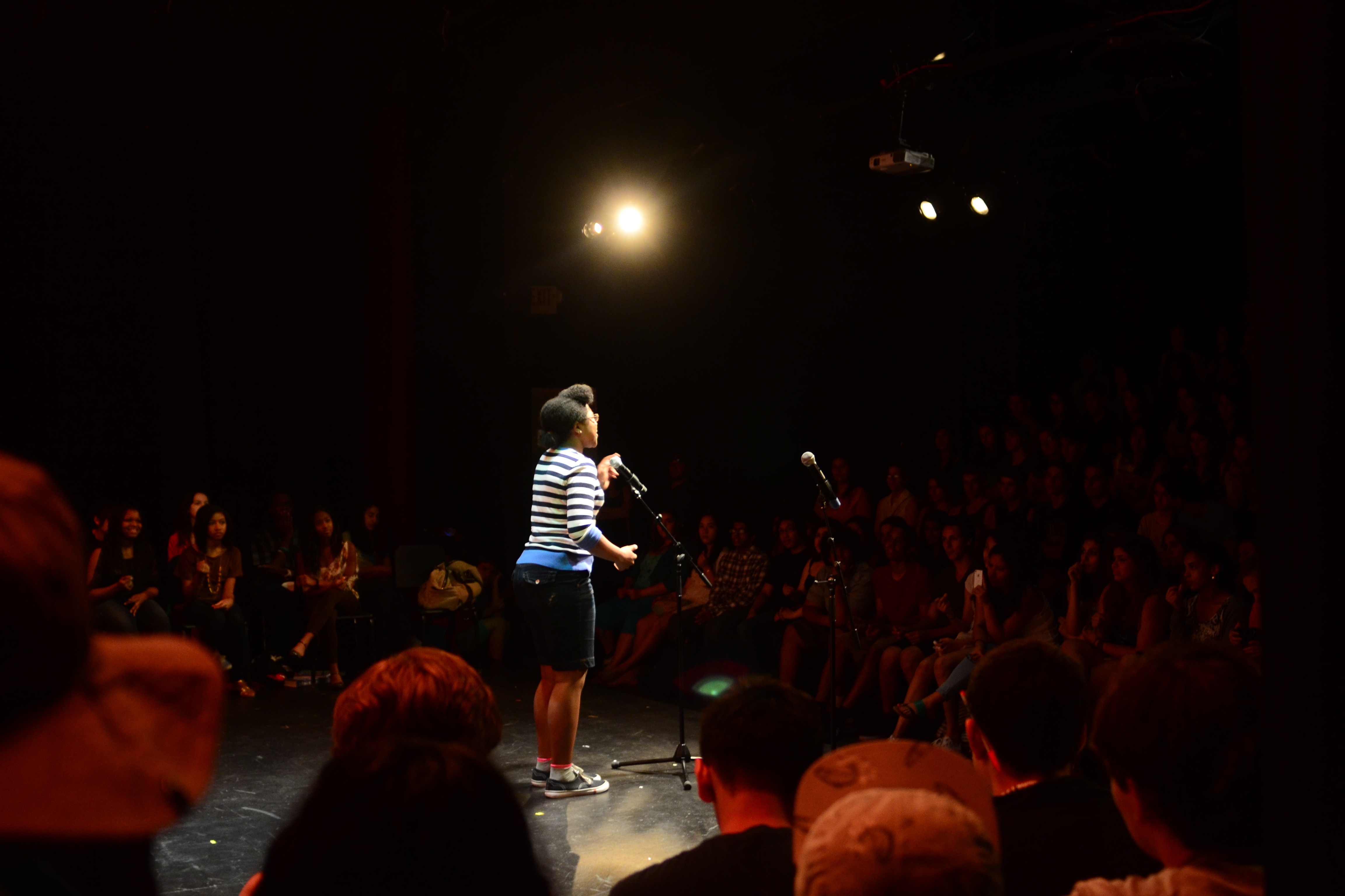A spotlit person wearing black shorts and a blue, white, and black sweater performs at a microphone on a stage